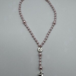 May include: A rosary necklace with light pink beads of varying sizes. The necklace features a silver-toned centerpiece with a religious image and a silver cross pendant at the bottom. The beads are strung together to form a loop.
