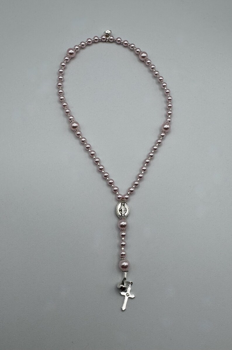 May include: A rosary necklace with light pink beads of varying sizes. The necklace features a silver-toned centerpiece with a religious image and a silver cross pendant at the bottom. The beads are strung together to form a loop.