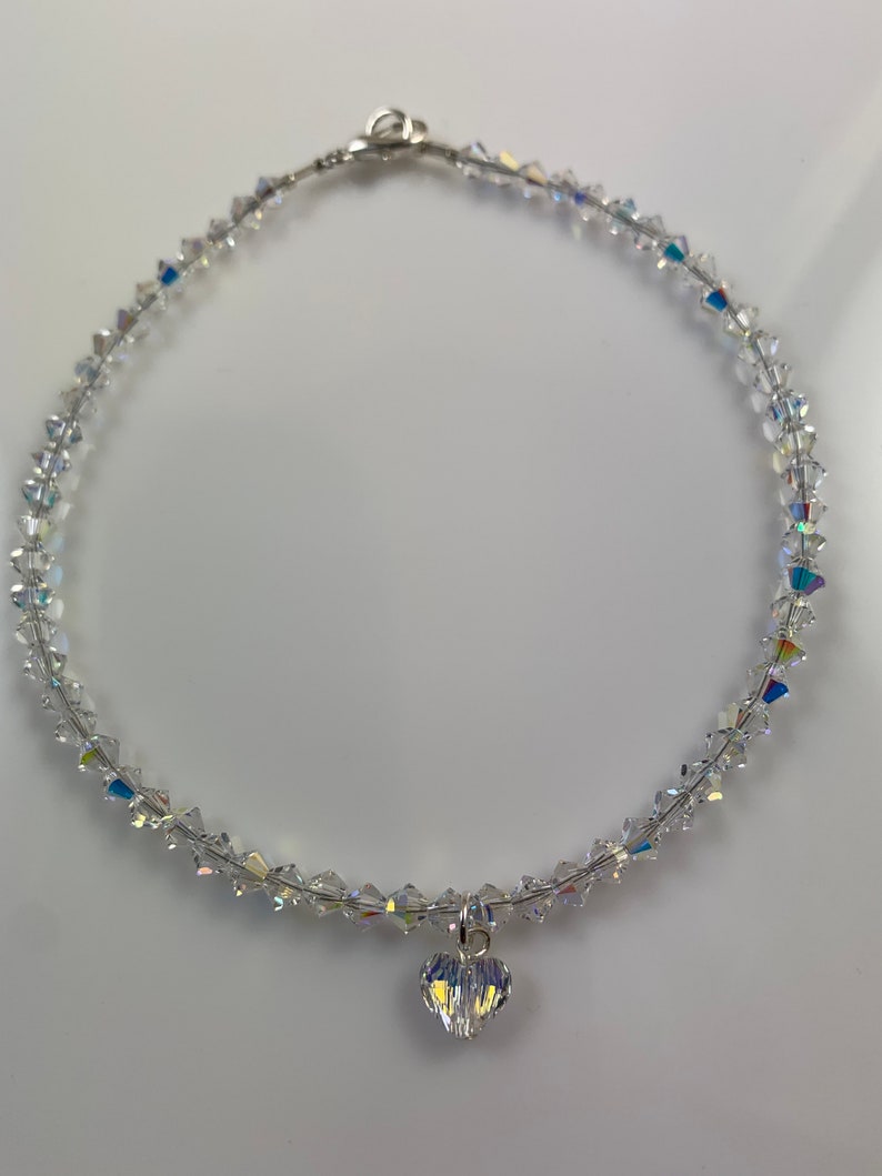 Pu&ograve; includere: Una collana di cristallo trasparente con un ciondolo a forma di cuore. La collana &egrave; composta da piccoli cristalli sfaccettati infilati su una sottile catena color argento. Il ciondolo a forma di cuore &egrave; anch'esso realizzato in cristalli trasparenti. La collana &egrave; esposta su una superficie bianca.
