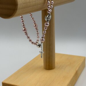 May include: A pink rosary with pearl beads and a silver crucifix. The rosary is displayed on a wooden stand. The beads are a soft pink color, and the crucifix and centerpiece are silver.