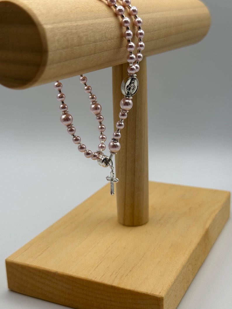 May include: A pink rosary with pearl beads and a silver crucifix. The rosary is displayed on a wooden stand. The beads are a soft pink color, and the crucifix and centerpiece are silver.