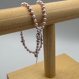 May include: A pink beaded rosary with a silver-tone centerpiece and crucifix. The rosary is displayed on a light brown wooden stand against a white background. The beads vary in size, creating a textured appearance.