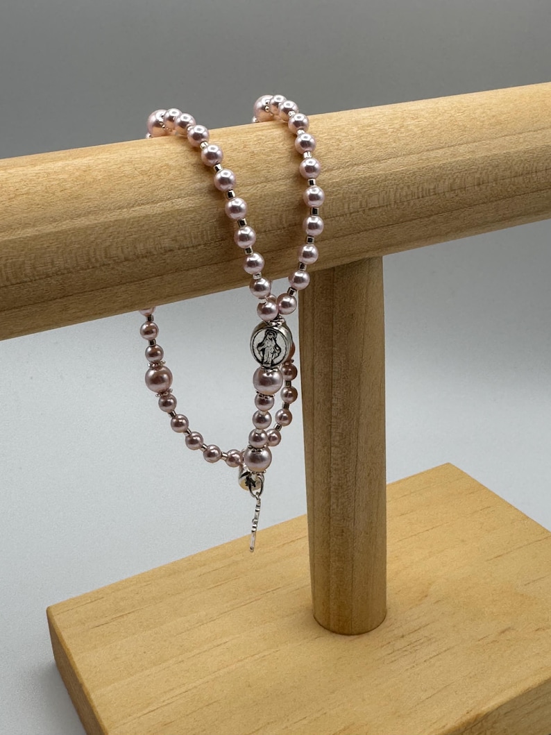 May include: A pink beaded rosary with a silver-tone centerpiece and crucifix. The rosary is displayed on a light brown wooden stand against a white background. The beads vary in size, creating a textured appearance.
