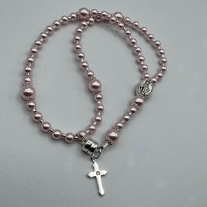 May include: A pink rosary necklace with a silver cross pendant. The necklace features round, pink beads of varying sizes. The cross has a small, clear gem in the center. The rosary is designed for religious use.
