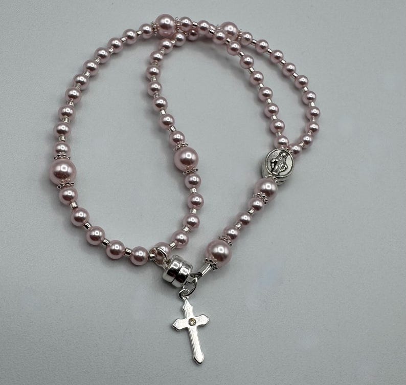 May include: A pink rosary necklace with a silver cross pendant. The necklace features round, pink beads of varying sizes. The cross has a small, clear gem in the center. The rosary is designed for religious use.