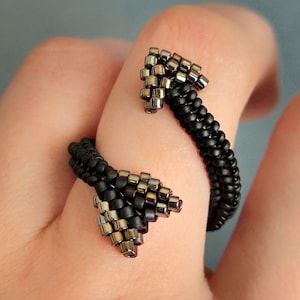 May include: A black beaded ring with silver accents. The ring features an open design with two arrow-shaped ends. The beads are small and create a textured surface. The ring is worn on a finger.