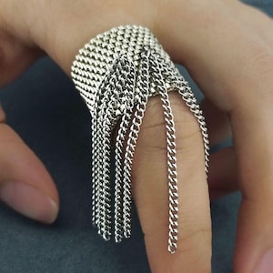 May include: Silver-toned ring featuring a wide band with a woven texture and multiple dangling chain strands. The ring is worn on a finger, showcasing its intricate design and metallic sheen. The jewelry piece has a modern, edgy aesthetic.
