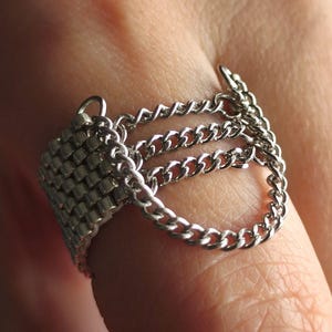 Może przedstawiać: Srebrny pierścionek z tkanym kwadratowym wzorem i wieloma zwisającymi łańcuszkami. Pierścionek jest noszony na palcu, prezentując jego skomplikowane detale łańcuszka i metaliczny połysk. Biżuteria ma unikalną, odważną estetykę.