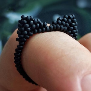 Könnte beinhalten: Ein schwarzer Perlenring in Form einer Fliege. Der Ring besteht aus kleinen, runden Perlen und wird an einem Finger getragen. Der Hintergrund ist verschwommen, mit Grün- und Brauntönen.