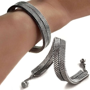 Puede incluir: Una pulsera plateada con un diseño de malla tejida. La pulsera se muestra en una muñeca y también por separado, mostrando su construcción flexible y su cierre magnético. La pulsera de metal tiene una estética elegante y moderna.