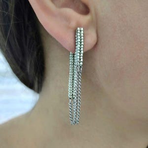 May include: Silver-toned dangle earrings. The earrings feature a rectangular post with a beaded design, from which multiple strands of delicate chain hang. The earrings are shown worn on an ear.