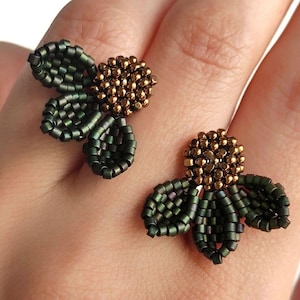 Pode incluir: Um par de brincos de contas feitos à mão com um design floral. Os brincos apresentam folhas verdes escuras e um aglomerado de contas douradas no centro. Os brincos são mostrados em uma mão.