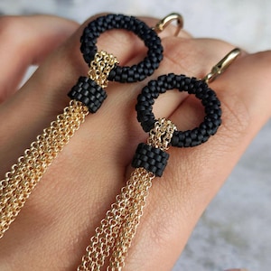 Puede incluir: Pendientes de aro negros con cuentas y borlas de cadena dorada. Los pendientes presentan un diseño circular con una textura de cuentas y una cadena dorada que cuelga. Diseñados como un accesorio de moda.