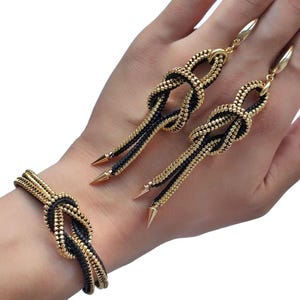 Puede incluir: Un conjunto de joyas de cremallera doradas y negras, que incluye una pulsera y pendientes. La pulsera está anudada y los pendientes cuelgan. El conjunto tiene un diseño único con metal dorado y tela negra.