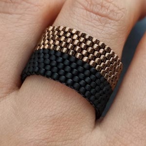 Może przedstawiać: Ręcznie robiony pierścionek z koralików o dwukolorowym wzorze. Pierścionek podzielony jest na dwie sekcje: czarny dolny pasek i miedziany górny pasek. Pierścionek wykonany jest z małych, równomiernie rozmieszczonych koralików, tworzących teksturowaną powierzchnię.