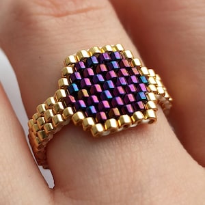Könnte beinhalten: Ein handgefertigter Perlenring mit einem quadratischen, irisierenden lila und blauen Mittelteil. Das Band des Rings und der Rand der Mitte sind goldfarben. Der Ring wird an einem Finger getragen.