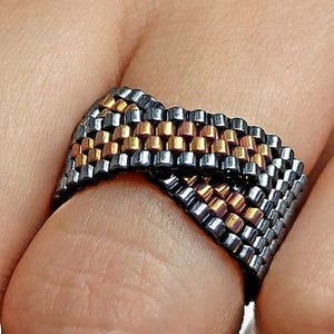 Könnte beinhalten: Ein gewebter Perlenring mit einem Muster aus metallischen Farben. Der Ring hat eine dunkelgraue Basis mit goldenen und bronzefarbenen Akzenten. Der Ring wird am Finger getragen.