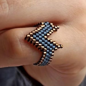 May include: A beaded ring in a chevron design. The ring features a pattern of small, light blue and bronze beads. The ring is worn on a finger, showcasing the intricate beadwork and geometric shape. The ring's design is a modern take on a classic accessory.