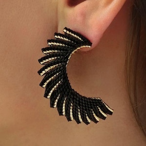 Può includere: Primo piano di un orecchino nero e oro. L'orecchino ha una forma a semicerchio con un design a strati. Gli strati sono neri con accenti dorati. L'orecchino è indossato sull'orecchio di una persona.