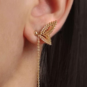 Puede incluir: Pendiente de color dorado con un diseño de ala de mariposa que se coloca en la oreja, con una delicada cadena que cuelga. El pendiente tiene una pequeña bola al final de la cadena. La joya se lleva en la oreja.