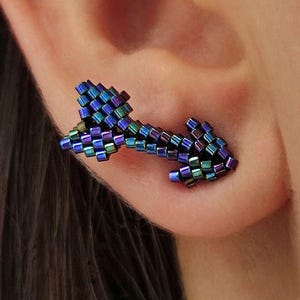 Puede incluir: Un pendiente en forma de flecha, hecho de pequeñas cuentas cuadradas en tonos azules, verdes y morados. El pendiente se muestra en una oreja. Un accesorio único y llamativo, ideal para añadir un toque de originalidad.