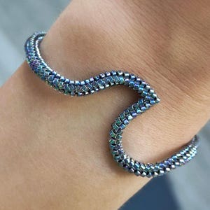 May include: A beaded bracelet in shades of blue, green, and purple, with a wavy design. The bracelet is made of small, iridescent beads and is worn on a wrist.