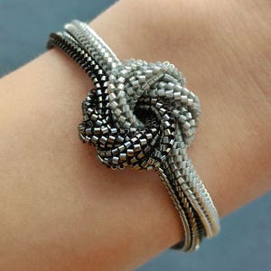 May include: A beaded bracelet with a knot design. The bracelet features a combination of silver and black beads, creating a two-tone effect. The knot is the focal point, with the bracelet's band wrapping around the wrist.