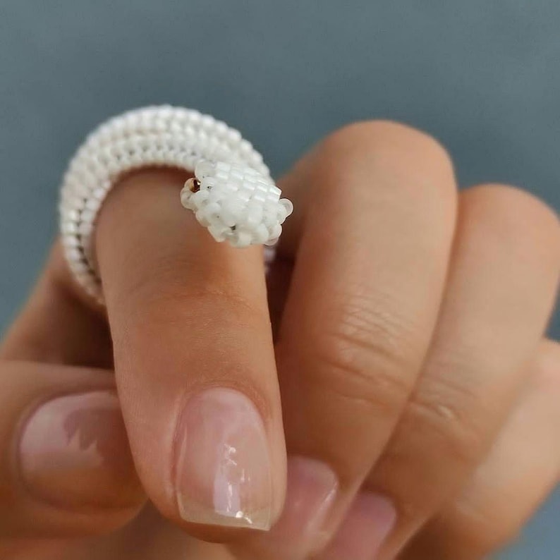 Puede incluir: Un anillo de serpiente blanco con cuentas enrollado alrededor de un dedo. El anillo est&aacute; hecho de peque&ntilde;as cuentas blancas redondas, con una cabeza de serpiente detallada y peque&ntilde;os ojos marrones. Una joya &uacute;nica.