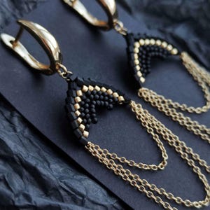 Könnte beinhalten: Nahaufnahme eines Paares gold-schwarzer Perlenohrringe. Die Ohrringe haben einen goldenen Bügel, ein schwarz-goldenes Perlendreieck und mehrere goldene Kettentrossen. Die Ohrringe werden auf einer dunklen Oberfläche präsentiert.