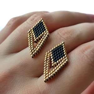 May include: A pair of diamond-shaped earrings crafted from small, cylindrical beads. The design features a central black bead pattern surrounded by a metallic gold bead border, creating an open, geometric structure with a distinct pointed shape.