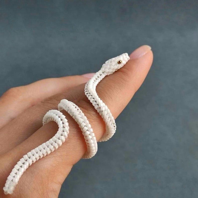 Puede incluir: Un anillo de serpiente blanco con cuentas enrollado alrededor de un dedo. El anillo presenta un dise&ntilde;o detallado con una cabeza de serpiente y un cuerpo enrollado. El ojo de la serpiente es una peque&ntilde;a cuenta dorada. El fondo es de color verde azulado apagado.