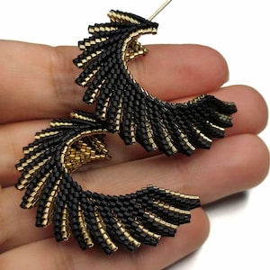 May include: A pair of handmade earrings featuring a fan-like design. The earrings are crafted with small black and gold beads, creating a textured, layered effect. The gold beads accent the edges and inner details of the black beaded design.