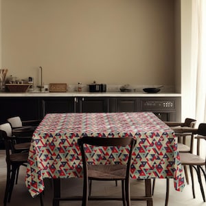 May include: A rectangular tablecloth featuring a repeating geometric pattern of red, blue, and beige triangles. The tablecloth is on a wooden dining table, surrounded by wooden chairs. The room has a neutral colour palette.