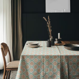 Puede incluir: Una mesa de comedor con un mantel estampado en verde claro y melocotón. La mesa está puesta con platos, un candelabro y un jarrón decorativo. Una silla de madera está colocada a la izquierda de la mesa. El fondo presenta una pared oscura y una ventana con cortinas.