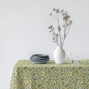 May include: A light green floral tablecloth featuring a repeating pattern of green vines and blue, yellow, and purple flowers. A stack of grey plates, a white vase with dried flowers, and two glasses of water are on the table.
