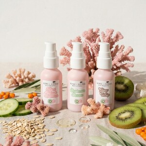 Three pink skincare bottles from Kiwi + Coral Skincare arranged in a row on a clean, bright background. The trio includes Kiwi Splash Cleanser, Sunbeam Serum, and Glow Whip, accented by natural props like sliced kiwi and decorative coral pieces to highlight the brand identity