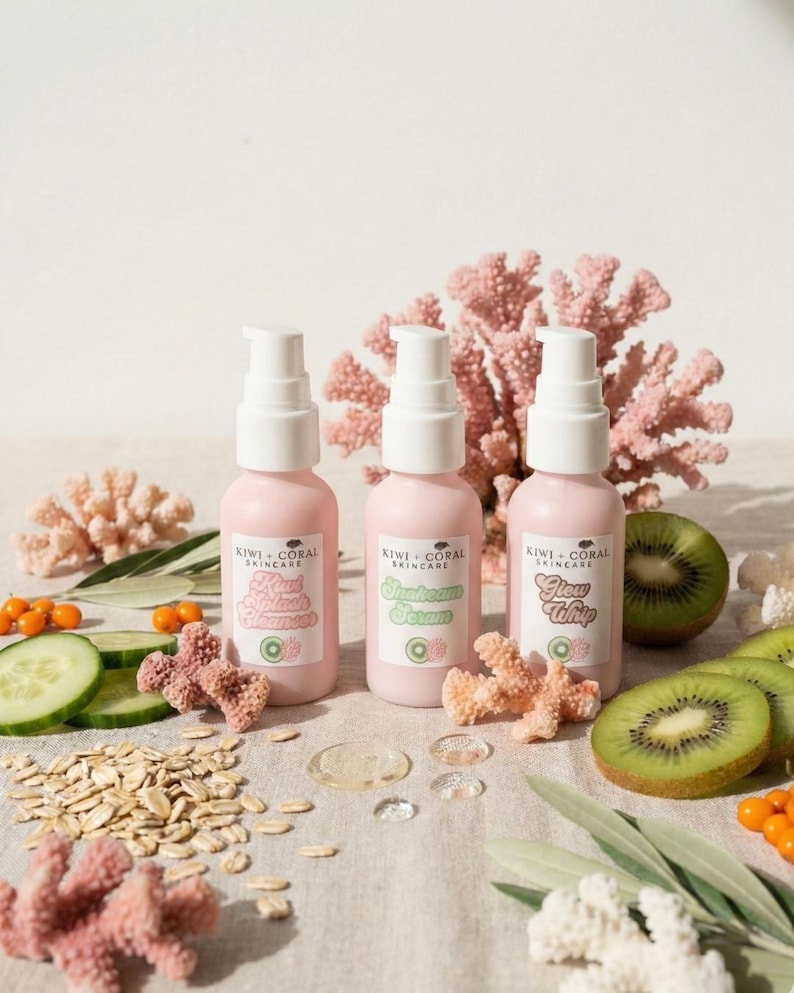 Three pink skincare bottles from Kiwi + Coral Skincare arranged in a row on a clean, bright background. The trio includes Kiwi Splash Cleanser, Sunbeam Serum, and Glow Whip, accented by natural props like sliced kiwi and decorative coral pieces to highlight the brand identity