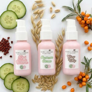 Three pink KIWI + CORAL skincare bottles, including Kiwi Splash Cleanser, Sunbeam Serum, and Glow Whip, laid flat on a white background. The bottles are surrounded by natural ingredients such as fresh cucumber slices, oat stalks and grains, sea buckthorn berries with leaves, and annatto seeds, showcasing the botanical components of the products.