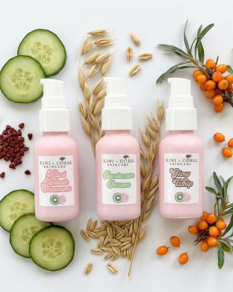 Three pink KIWI + CORAL skincare bottles, including Kiwi Splash Cleanser, Sunbeam Serum, and Glow Whip, laid flat on a white background. The bottles are surrounded by natural ingredients such as fresh cucumber slices, oat stalks and grains, sea buckthorn berries with leaves, and annatto seeds, showcasing the botanical components of the products.