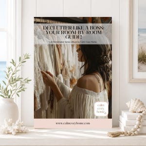 以下が含まれることがあります： 電子書籍の表紙。タイトルは「DECLUTTER LIKE A BOSS: YOUR ROOM-BY-ROOM GUIDE! A Homemaker Series eBook by Calm Cozy Home」。白いオフショルダーのトップスを着た人物が、白い服が並ぶハンガーラックを見ている様子が写っています。ウェブサイト: www.calmcozyhome.com。