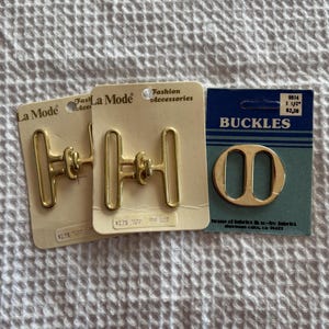 May include: Three fashion accessory buckles. Two gold-colored metal H-shaped buckles on cardboard backing labeled "La Mode" and "Fashion Accessories". A third gold-colored oval buckle is on a blue card labeled "BUCKLES".