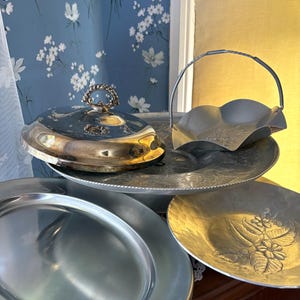 May include: A collection of vintage silver serving dishes and trays. The set includes an oval platter, a covered dish with a decorative handle, a handled basket-shaped dish, and a round tray with floral detailing. The pieces are arranged on a wooden surface.