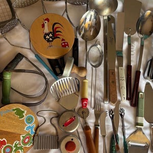 May include: A collection of vintage kitchen utensils, including spatulas, ladles, and mashers. A wooden cutting board with a rooster design is also present. The utensils are made of metal and wood, with various handle colors and styles. The items are arranged on a white surface.