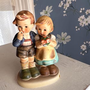 May include: A ceramic figurine depicting a boy and girl in traditional attire. The boy wears a blue jacket, brown shorts, and green shoes, while the girl has a dress with a yellow apron and holds a bouquet. The base is oval-shaped.