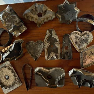 May include: A collection of vintage metal cookie cutters in various shapes, including a bat, heart, star, and geometric designs. The cutters are made of tarnished metal and are arranged on a wooden surface. The collection includes a variety of sizes and styles.