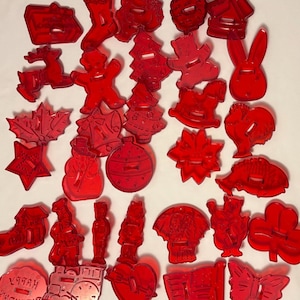 May include: A collection of red translucent plastic cookie cutters in various shapes including a Christmas tree, reindeer, star, heart, butterfly, windmill, and a round shape with the text Happy Birthday embossed on the surface of the plastic.