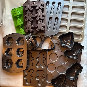 May include: A collection of various baking molds and cookie cutters. Includes gingerbread man, Christmas tree, heart, and circle shapes. The molds are made of metal and silicone in shades of brown, green, and silver.