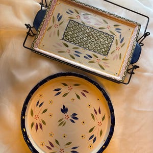 May include: Two ceramic serving dishes. One is rectangular with blue handles and a floral design in green, pink, and blue. The other is round with a blue rim and a similar floral pattern. Both are on a white surface.