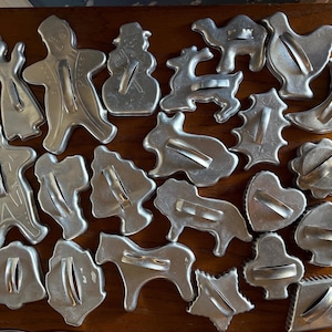 May include: A collection of twenty-two silver-colored metal cookie cutters in various shapes, including figures of people, animals, holiday symbols, and geometric forms. Each cutter features a central metal handle for gripping and pressing.