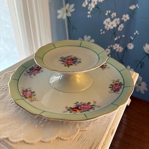 May include: A two-tiered, vintage-style cake stand. The stand features a light green border with a gold trim and floral designs. The stand is made of ceramic and is placed on a white surface.